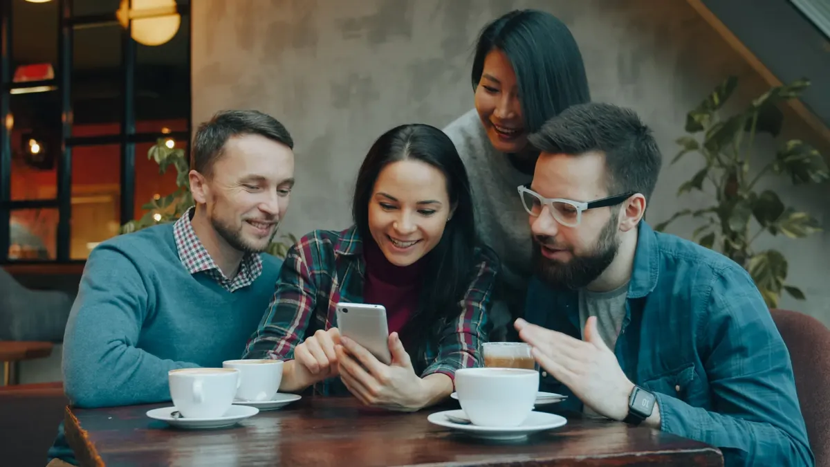 Four friends sharing a moment over coffee in a cozy cafe setting, laughing and engaging with a smartphone.