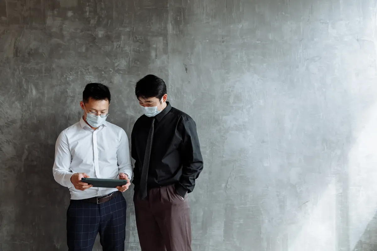 Two businessmen wearing face masks use a digital tablet against a modern gray background.
