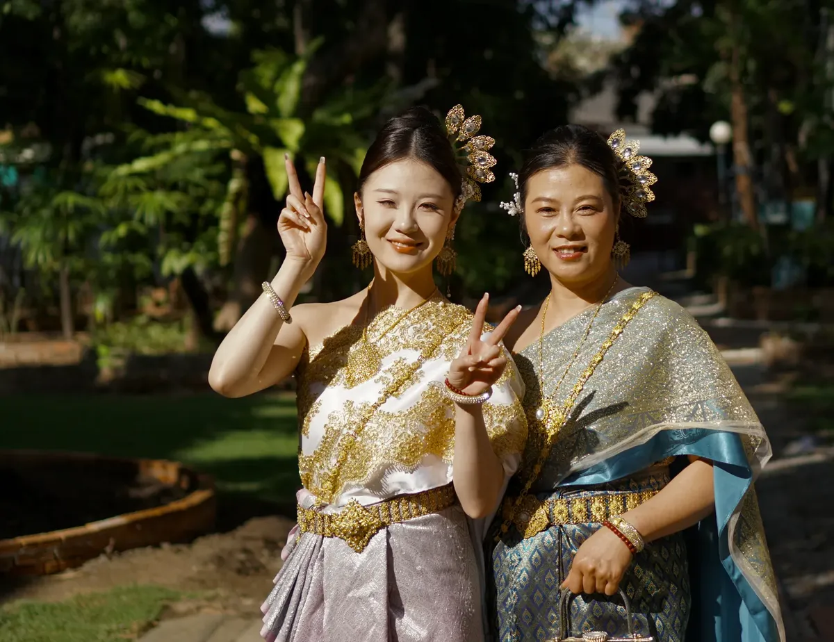 Two smiling women in traditional Thai costumes enjoying a sunny day outdoors.